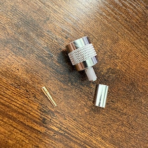 May include: A disassembled silver-colored RF connector with a knurled grip, a small gold-colored pin, and a silver-colored sleeve, all resting on a wooden surface. The connector is designed for radio frequency applications.