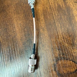 May include: A silver-colored RF connector cable with a beige cable and black heat shrink tubing. The connectors have a ribbed design for secure grip. The cable is designed for connecting radio frequency devices.