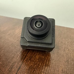 May include: A small, square, dark gray camera lens with a circular black lens. The lens is mounted on a dark gray base. The camera is sitting on a brown wooden surface.