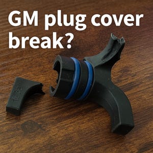 Puede incluir: Una cubierta de enchufe GM negra rota con detalles azules. La cubierta está en dos piezas, con el texto "GM plug cover break?" en blanco encima del objeto. Sobre una superficie de madera marrón.