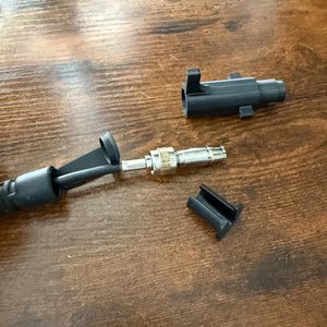 Pode incluir: Um conector elétrico preto desmontado com um componente metálico prateado e uma peça de plástico preta separada. Os componentes estão dispostos sobre uma superfície de madeira.