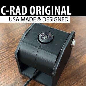 Może przedstawiać: Czarna kamera C-RAD ORIGINAL z obiektywem i kablem. Tekst "USA MADE & DESIGNED" znajduje się nad kamerą. Kamera znajduje się na drewnianej powierzchni.