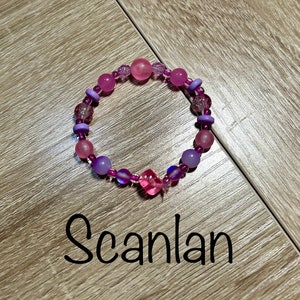 May include: A purple and pink beaded bracelet with a pink square charm. The bracelet is on a wooden surface with the word "Scanlan" written below it.