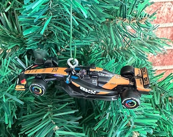 May include: A miniature Formula 1 race car ornament featuring a black and orange color scheme with the number 81 and various sponsor logos including OKX, Chrome, and Dell printed on the body, suspended by a silver metal loop for hanging.