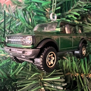 May include: A green Bronco SUV Christmas ornament. The miniature vehicle has black tires with gold rims and a silver front grill with the word "BRONCO" across the front. The ornament hangs from a green string.