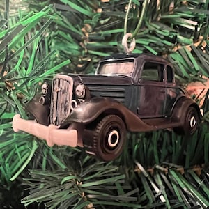 May include: A dark blue vintage car ornament, hanging from a green artificial Christmas tree. The car has a chrome bumper and headlights.