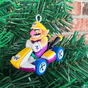 May include: A Christmas tree ornament featuring Wario in a yellow kart. The kart has blue wheels and purple accents. Wario is wearing a yellow hat and purple overalls. The ornament is hanging from a green string.