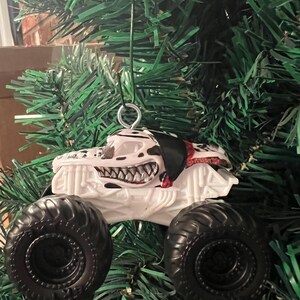 May include: A white monster truck Christmas ornament with black tires and a Dalmatian-themed design. The truck features a menacing grin and black and white spots. The ornament hangs from a silver metal loop.