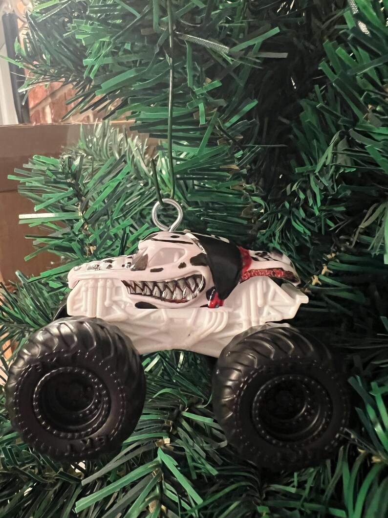 May include: A white monster truck Christmas ornament with black tires and a Dalmatian-themed design. The truck features a menacing grin and black and white spots. The ornament hangs from a silver metal loop.