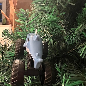 May include: A light blue monster truck ornament shaped like a shark, with black wheels, hangs on a green Christmas tree. The shark truck has a silver hook for hanging. The ornament is a fun, festive decoration.