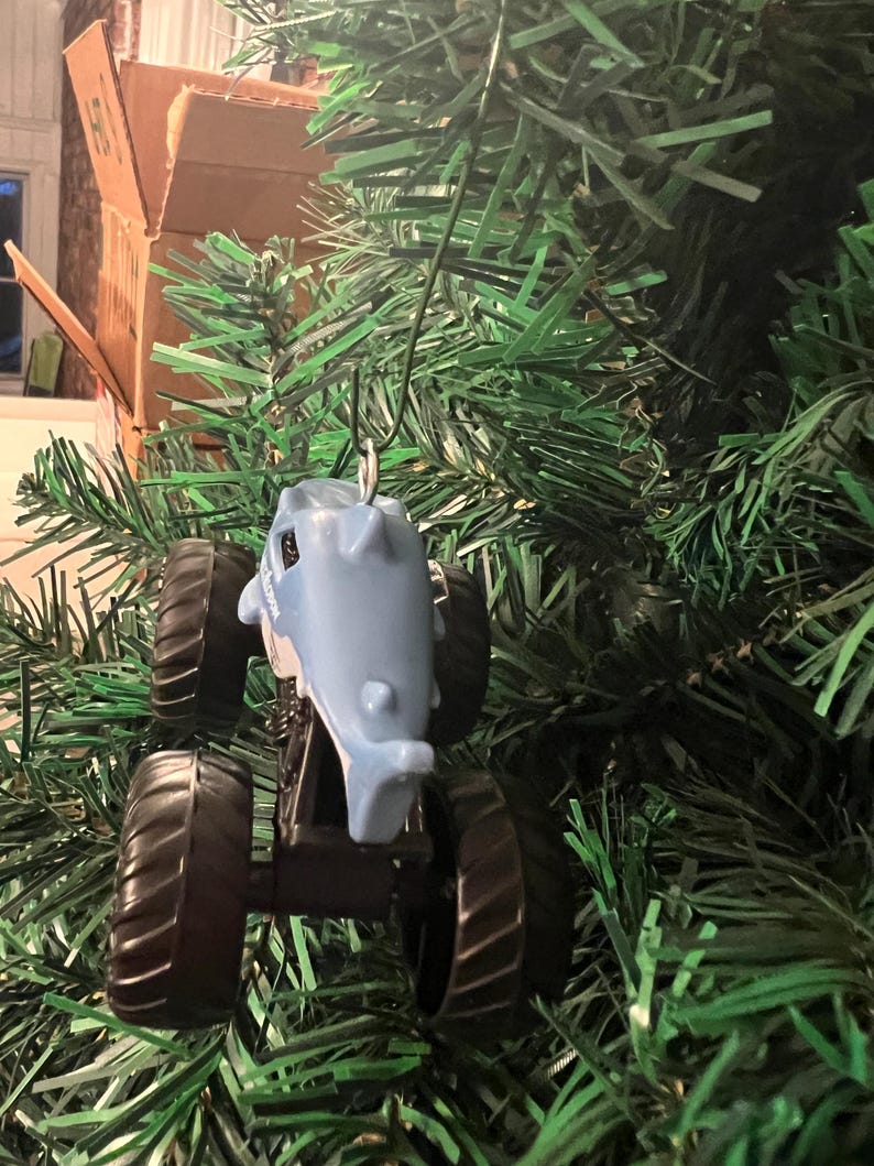 May include: A light blue monster truck ornament shaped like a shark, with black wheels, hangs on a green Christmas tree. The shark truck has a silver hook for hanging. The ornament is a fun, festive decoration.
