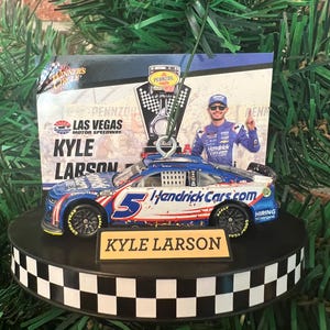 May include: A Christmas tree ornament featuring a miniature race car with the number 5 and the name "Kyle Larson." The ornament has a black and white checkered base and a backdrop with the words "Las Vegas Motor Speedway."