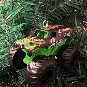 May include: A Grave Digger monster truck ornament with black tires, a green chassis, and a black cab with green and orange flames. The truck has the words "Grave Digger" on the side and is hanging on a Christmas tree.