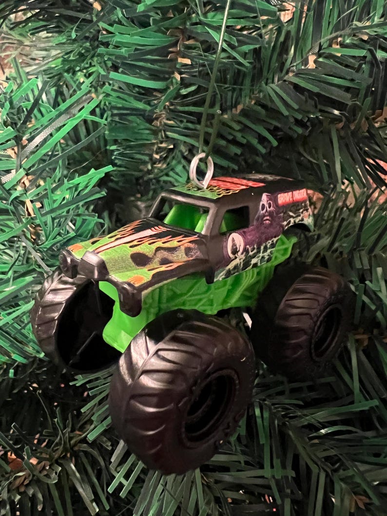 May include: A Grave Digger monster truck ornament with black tires, a green chassis, and a black cab with green and orange flames. The truck has the words "Grave Digger" on the side and is hanging on a Christmas tree.
