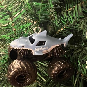 May include: A light blue monster truck ornament shaped like a shark, with the word "Megalodon" printed on the side. The truck has large black tires and is hanging from a silver hook against a backdrop of green Christmas tree branches.