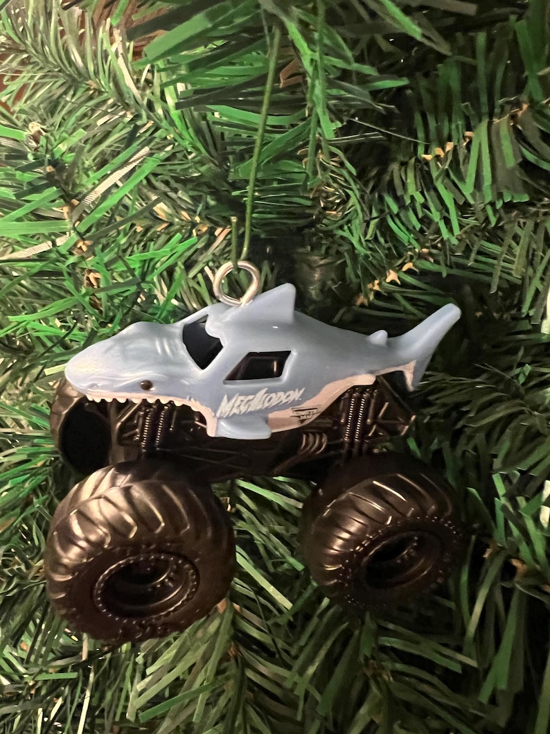 May include: A light blue monster truck ornament shaped like a shark, with the word "Megalodon" printed on the side. The truck has large black tires and is hanging from a silver hook against a backdrop of green Christmas tree branches.