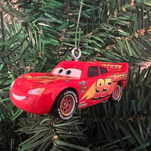May include: A red Lightning McQueen car ornament with the number 95 on the side. The ornament has yellow and orange flames and the word "RUST-EZE" on the back. The ornament is hanging on a green Christmas tree.