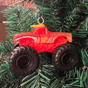 Puede incluir: Adorno navide&ntilde;o de cami&oacute;n monstruo rojo con neum&aacute;ticos negros y el texto "El Toro Loco". El cami&oacute;n tiene un techo amarillo y un dise&ntilde;o de cara sonriente. Cuelga de un &aacute;rbol de Navidad verde.