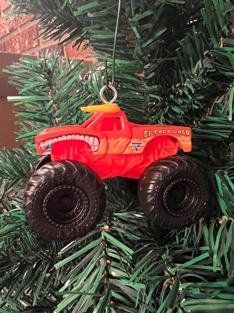 Puede incluir: Adorno navide&ntilde;o de cami&oacute;n monstruo rojo con neum&aacute;ticos negros y el texto "El Toro Loco". El cami&oacute;n tiene un techo amarillo y un dise&ntilde;o de cara sonriente. Cuelga de un &aacute;rbol de Navidad verde.