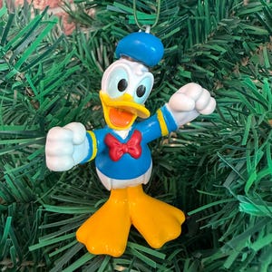 May include: A Donald Duck Christmas ornament featuring the character in a blue sailor shirt with a red bow tie, a blue cap, and yellow feet. The figure has white arms and a yellow beak, with a metal hook attached to the top of the blue cap.