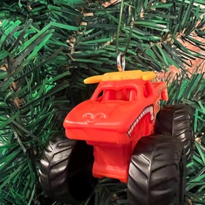 Puede incluir: Adorno navide&ntilde;o de cami&oacute;n monstruo naranja con neum&aacute;ticos negros y un aler&oacute;n amarillo. El cami&oacute;n tiene una cara sonriente y una boca con dientes blancos y negros. El adorno cuelga de una cuerda verde sobre un fondo de ramas verdes de &aacute;rbol de Navidad.