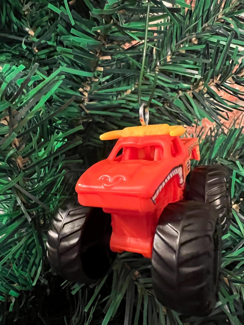 Puede incluir: Adorno navide&ntilde;o de cami&oacute;n monstruo naranja con neum&aacute;ticos negros y un aler&oacute;n amarillo. El cami&oacute;n tiene una cara sonriente y una boca con dientes blancos y negros. El adorno cuelga de una cuerda verde sobre un fondo de ramas verdes de &aacute;rbol de Navidad.