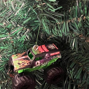 May include: A miniature Grave Digger monster truck ornament with a black body, green accents, and large black tires. The truck features flame and skull graphics, and the words "Grave Digger" are visible. The ornament hangs from a silver loop and rests on a bed of green Christmas tree needles.