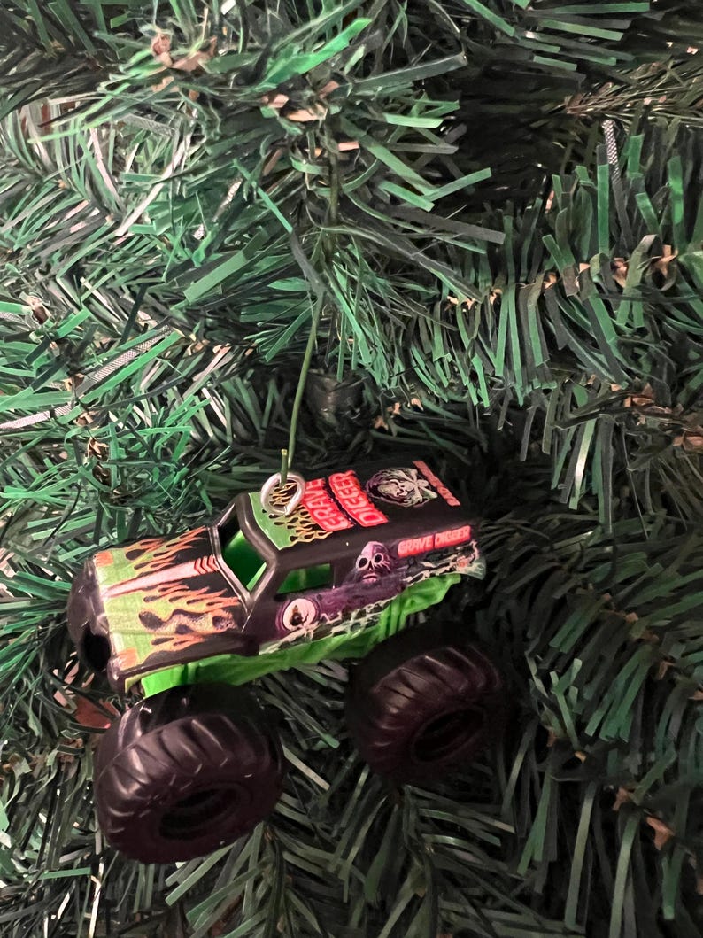 May include: A miniature Grave Digger monster truck ornament with a black body, green accents, and large black tires. The truck features flame and skull graphics, and the words "Grave Digger" are visible. The ornament hangs from a silver loop and rests on a bed of green Christmas tree needles.