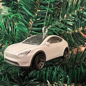 May include: A white Tesla Model X toy car Christmas ornament. The miniature vehicle has a black roof, bronze-colored wheels, and is suspended by a silver wire loop. The ornament is set against a backdrop of green Christmas tree branches.