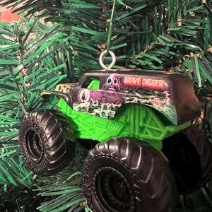May include: A Grave Digger monster truck ornament with black tires and a green chassis. The truck has a black cab with the words "Grave Digger" and a spooky graphic. The ornament is hanging on a green Christmas tree.