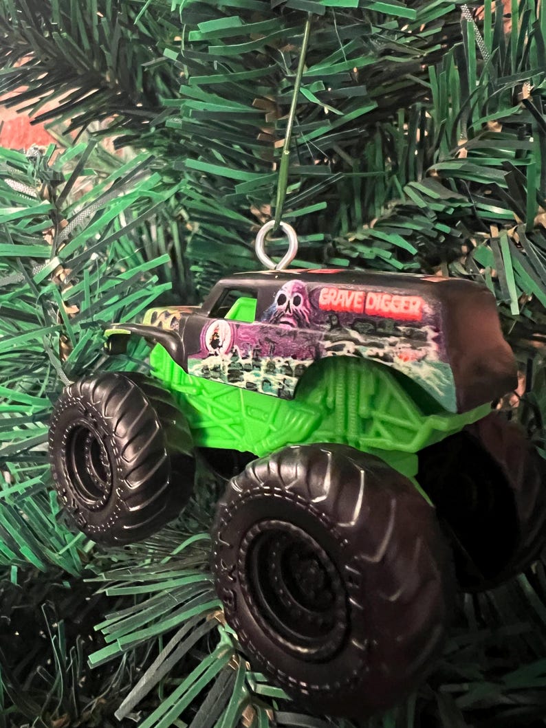 May include: A Grave Digger monster truck ornament with black tires and a green chassis. The truck has a black cab with the words "Grave Digger" and a spooky graphic. The ornament is hanging on a green Christmas tree.