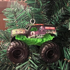 May include: A miniature Grave Digger monster truck ornament. The truck has a black cab with a green chassis and large black tires. The cab features a monster graphic and the words "Grave Digger". The ornament hangs from a silver metal loop.