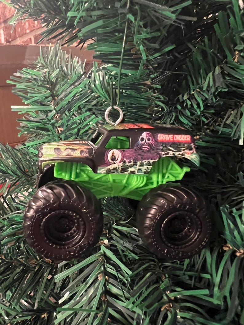 May include: A miniature Grave Digger monster truck ornament. The truck has a black cab with a green chassis and large black tires. The cab features a monster graphic and the words "Grave Digger". The ornament hangs from a silver metal loop.