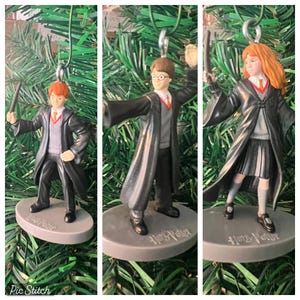 May include: Three Harry Potter character ornaments. Each figure is wearing a black robe and holding a wand. The characters are standing on a gray base with the words "Harry Potter" printed on it. The ornaments are hanging on a green Christmas tree.