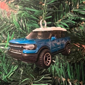 May include: A blue and white toy SUV Christmas ornament. The ornament has black tires and the word "BRONCO" is visible on the front. The ornament is hanging from a green string.