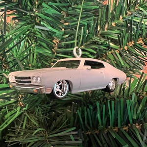 May include: A light gray classic car ornament with chrome wheels and trim. The car is suspended by a silver wire, ready for display. The ornament is set against a backdrop of green Christmas tree branches.