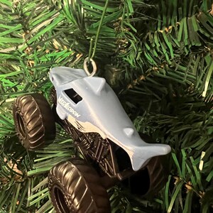 May include: A light blue toy monster truck shaped like a shark, with the word "MEGALODON" printed on the side. The truck has large black tires and is hanging from a green string, set against a backdrop of green Christmas tree branches.