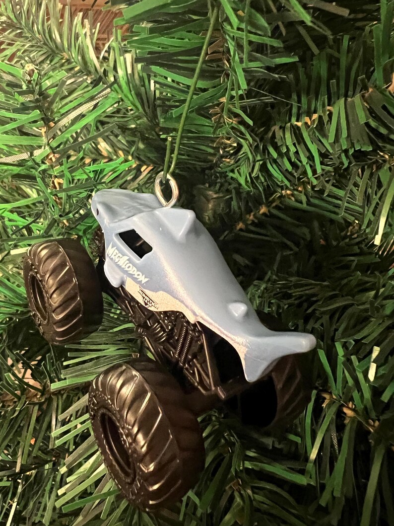 May include: A light blue toy monster truck shaped like a shark, with the word "MEGALODON" printed on the side. The truck has large black tires and is hanging from a green string, set against a backdrop of green Christmas tree branches.