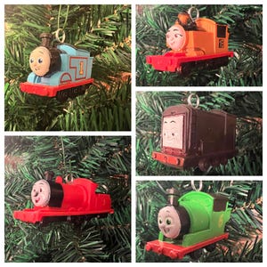 May include: A collage of five Christmas tree ornaments featuring cartoon train characters. The trains are in various colors, including blue, red, green, brown, and orange. Each train has a smiling face and a number on its side.