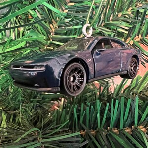 May include: A dark blue car ornament with black wheels and a silver hanging loop. The car has a sleek design and is suspended against a backdrop of green Christmas tree branches. A festive holiday decoration.