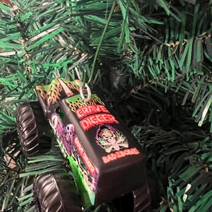 May include: A black Grave Digger monster truck ornament with a green and red flame design. The ornament has the words "Grave Digger" and "Bad Bone" printed on it. The ornament is hanging on a green Christmas tree.