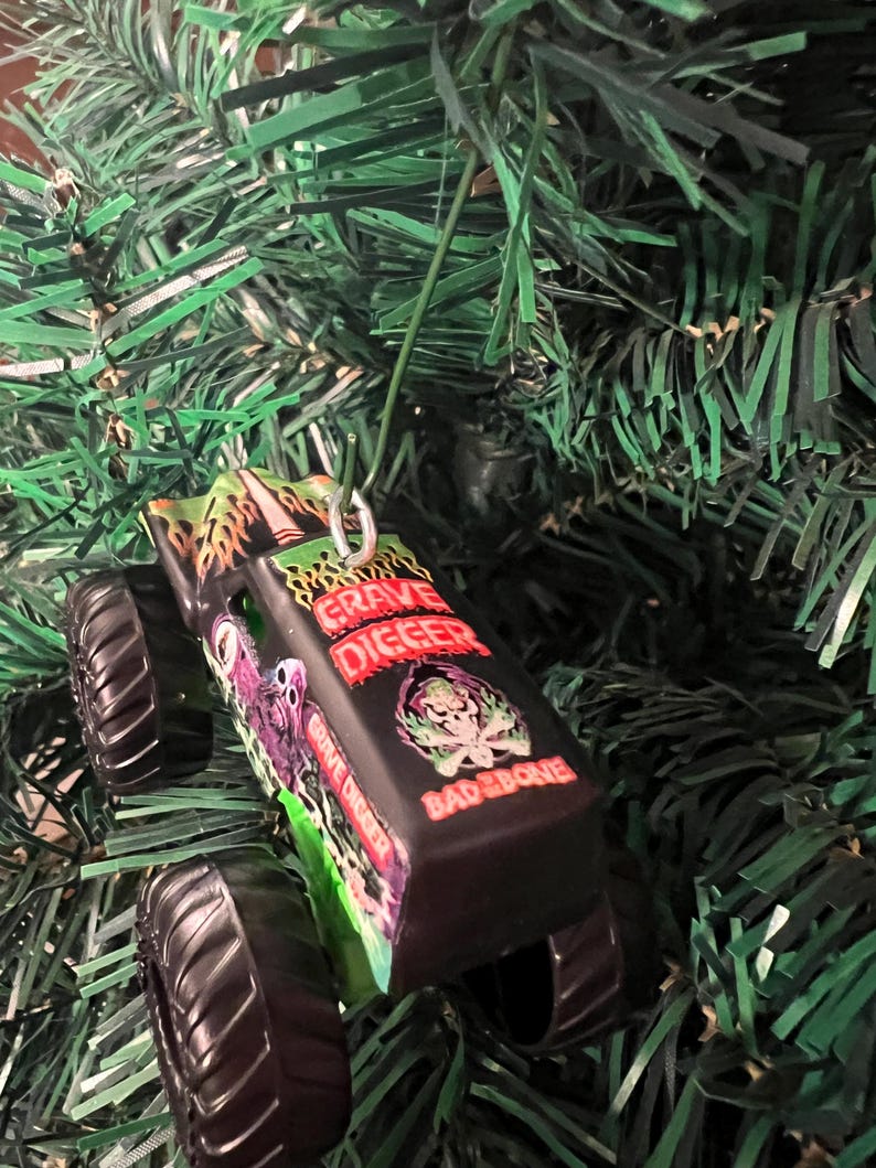 May include: A black Grave Digger monster truck ornament with a green and red flame design. The ornament has the words "Grave Digger" and "Bad Bone" printed on it. The ornament is hanging on a green Christmas tree.