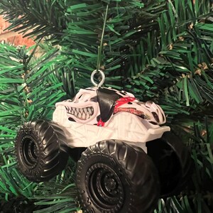 May include: White monster truck ornament with black tires and a menacing grin design. The truck has a black and red pattern on the top and is hanging from a green Christmas tree.