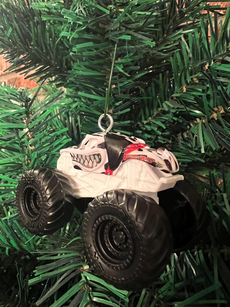 May include: White monster truck ornament with black tires and a menacing grin design. The truck has a black and red pattern on the top and is hanging from a green Christmas tree.