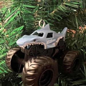 May include: A light blue and white Megalodon monster truck ornament with large black tires. The shark-themed truck has the word "MEGALODON" printed on the side. The ornament hangs from a silver metal loop and is set against a green Christmas tree.