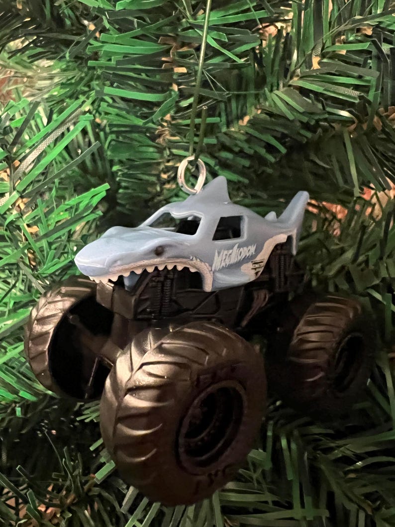 May include: A light blue and white Megalodon monster truck ornament with large black tires. The shark-themed truck has the word "MEGALODON" printed on the side. The ornament hangs from a silver metal loop and is set against a green Christmas tree.