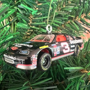 May include: A black die-cast race car ornament featuring the number 3, Goodwrench Service branding, and Monte Carlo text. The vehicle has black tires with Goodyear lettering, a red interior, and a silver metal loop attached to the roof for hanging.