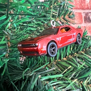May include: A red toy car Christmas ornament. The car has black wheels and a detailed design. The ornament is hanging from a silver hook and is set against a backdrop of green pine needles and a brick wall.
