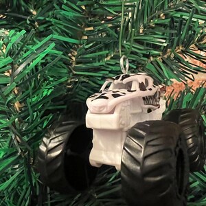 May include: A white and black monster truck Christmas ornament with large black tires. The truck has a dinosaur-like design on the front and is hanging from a green Christmas tree.