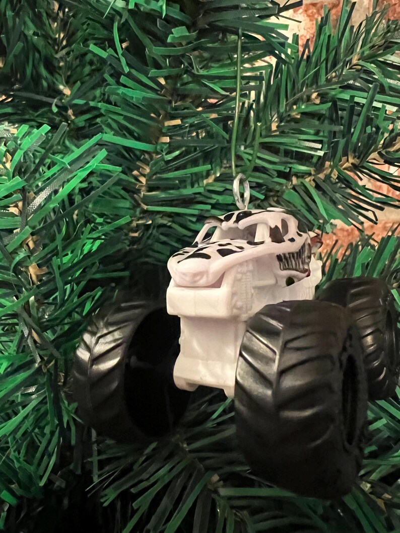 May include: A white and black monster truck Christmas ornament with large black tires. The truck has a dinosaur-like design on the front and is hanging from a green Christmas tree.